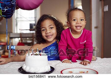 Stock Image - Mixed race sisters smiling with cake at party. Fotosearch