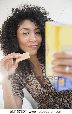 Stock Image - Mixed race woman eating cracker and reading nutrition information. Fotosearch