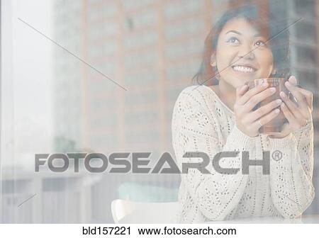 Stock Image - Pacific Islander woman drinking cup of coffee behind window. Fotosearch