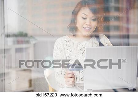 Stock Image - Pacific Islander woman shopping online on laptop behind window. Fotosearch