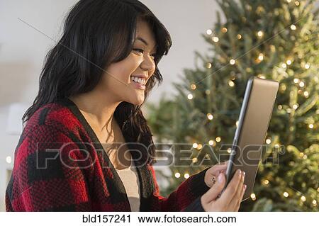 Stock Image - Pacific Islander woman using digital tablet near Christmas tree. Fotosearch