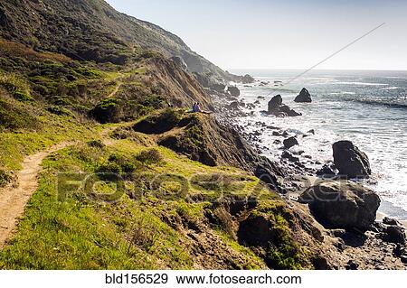 Stock Photo - People sitting on rocky coastline trail near ocean. Fotosearch