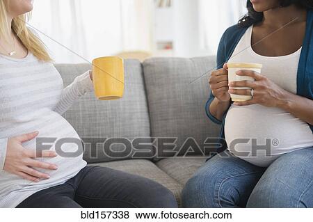 Stock Photo - Pregnant women drinking coffee and talking on sofa. Fotosearch