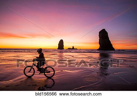 Stock Photograph - Silhouette of Caucasian girl biking near rock formations on Cannon Beach, Oregon, United States. Fotosearch
