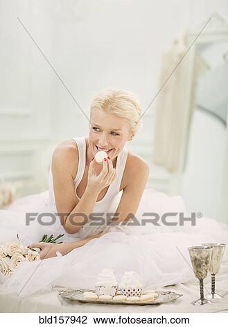 Stock Image - Smiling bride eating cupcakes on bed. Fotosearch