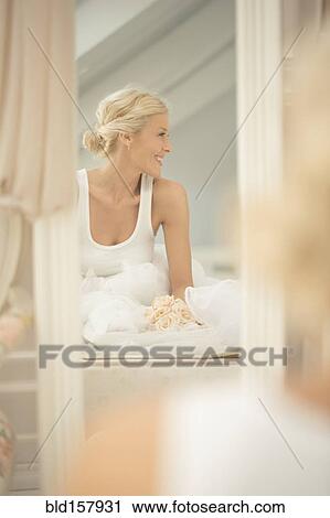 Stock Image - Smiling bride sitting on bed with bouquet in mirror. Fotosearch