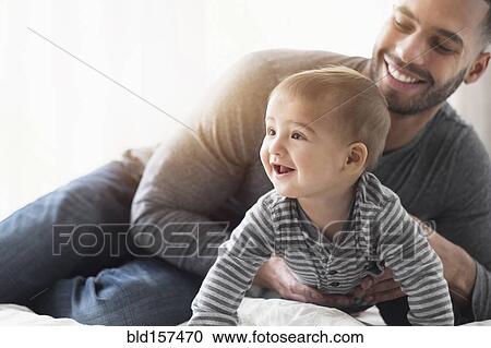 Smiling father playing with baby son on bed View Large Photo Image Stock Image - Smiling father playing with baby son on bed. Fotosearch
