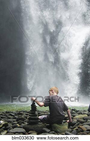Stock Image - Caucasian boy stacking rocks near waterfall. Fotosearch