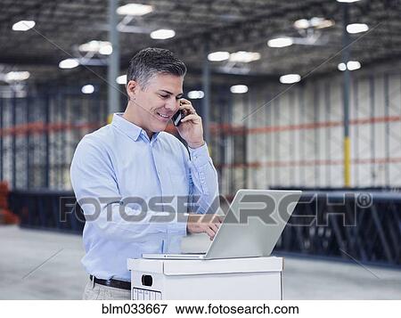 Stock Photo - Caucasian businessman using laptop and cell phone in empty warehouse. Fotosearch