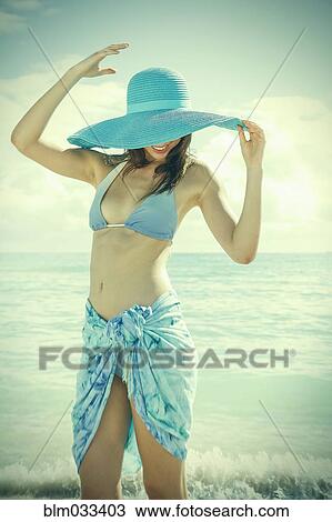 Stock Image - Caucasian woman wearing bikini and sun hat on beach. Fotosearch