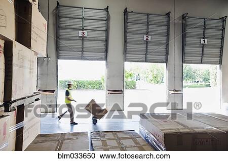 Stock Image - Caucasian worker wheeling cardboard boxes in warehouse. Fotosearch