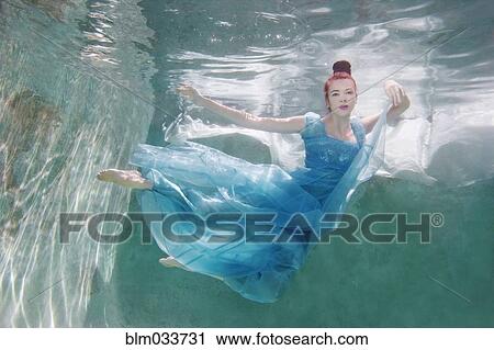 Stock Image - Underwater view of Caucasian woman in dress swimming in pool. Fotosearch