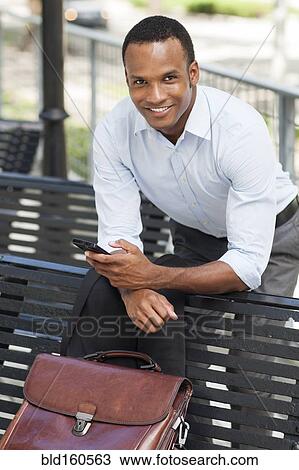 Stock Image - Black businessman using cell phone on park bench. Fotosearch