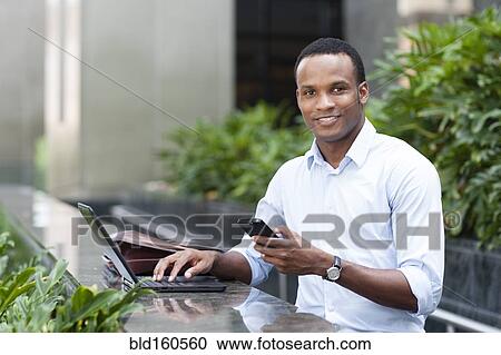 Stock Image - Black businessman using laptop and cell phone outdoors. Fotosearch