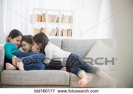 Stock Photo - Caucasian brother and sisters whispering on sofa. Fotosearch