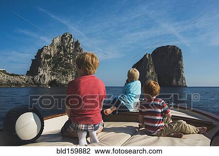 Stock Image - Caucasian brothers on boat admiring rock formations in ocean, Isle of Capri, Naples, Italy. Fotosearch