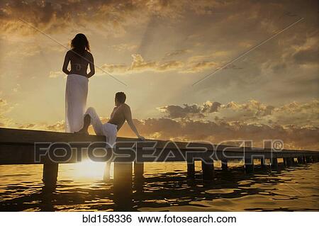 Stock Photograph - Caucasian couple admiring sunset from dock over ocean. Fotosearch