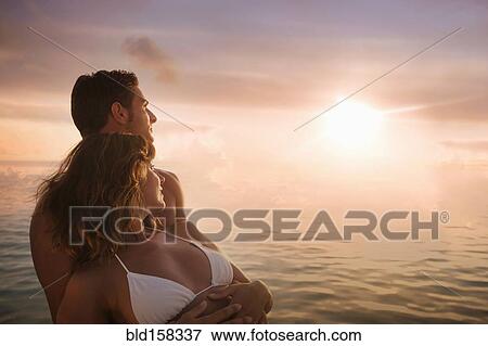 Stock Photo - Caucasian couple admiring sunset over ocean. Fotosearch