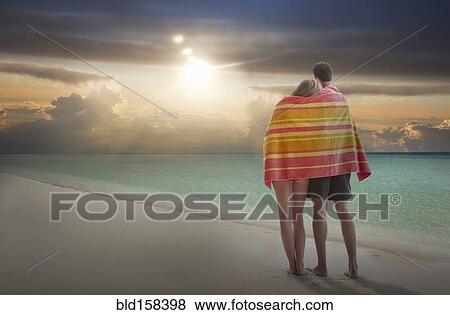 Stock Photo - Caucasian couple wrapped in towel admiring sunset on beach. Fotosearch