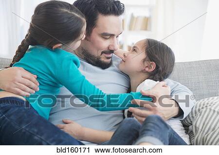 Stock Image - Caucasian father and daughters hugging on sofa. Fotosearch