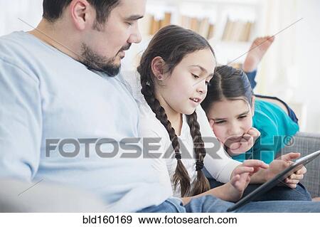 Stock Photo - Caucasian father and daughters using digital tablet on sofa. Fotosearch