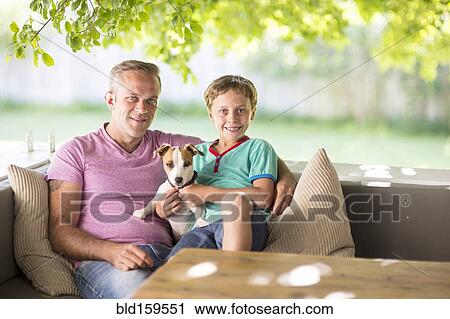 Stock Image - Caucasian father and son sitting with dog on sofa. Fotosearch