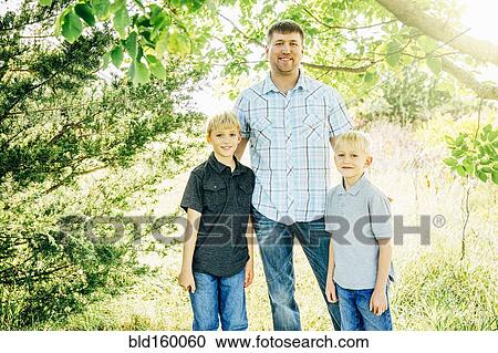 Stock Image - Caucasian father and sons smiling under trees. Fotosearch