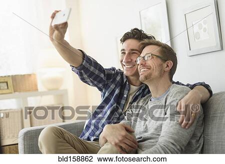 Stock Image - Caucasian gay couple taking selfie on sofa. Fotosearch