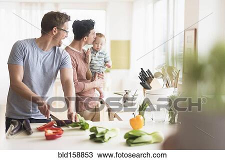 Stock Image - Caucasian gay fathers and baby cooking in kitchen. Fotosearch