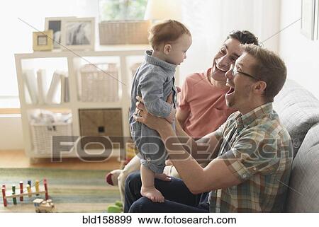 Stock Photo - Caucasian gay fathers and baby playing in living room. Fotosearch