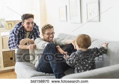 Stock Image - Caucasian gay fathers and baby relaxing on sofa. Fotosearch
