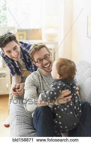 Stock Image - Caucasian gay fathers and baby relaxing on sofa. Fotosearch