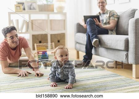Stock Image - Caucasian gay fathers and baby relaxing in living room. Fotosearch