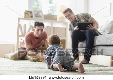 Stock Image - Caucasian gay fathers and baby relaxing in living room. Fotosearch