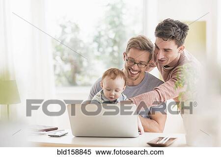 Stock Photography - Caucasian gay fathers and baby using laptop. Fotosearch