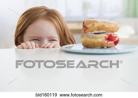 Stock Photo - Caucasian girl peeking at donuts on table. Fotosearch