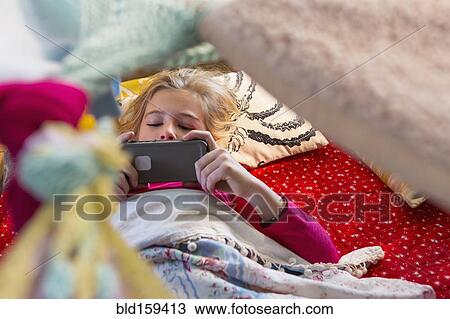 Stock Image - Caucasian girl using cell phone in blanket fort. Fotosearch