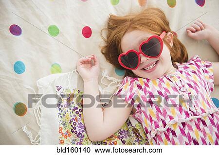 Stock Image - Caucasian girl wearing heart -shaped sunglasses on bed. Fotosearch