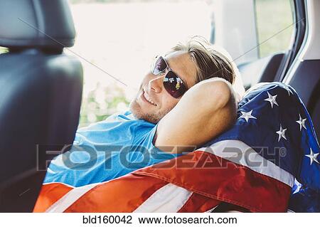Stock Image - Caucasian man laying on American flag in car. Fotosearch