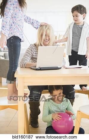 Stock Photo - Caucasian mother using laptop with chaotic children. Fotosearch