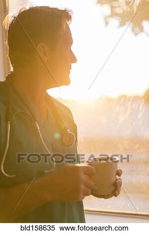 Stock Photography - Caucasian nurse drinking cup of coffee at window. Fotosearch