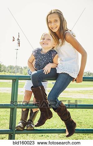 Stock Image - Caucasian sisters smiling on gates near farm field. Fotosearch