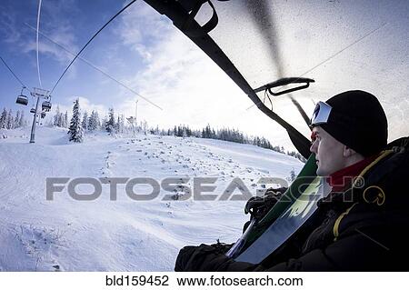Stock Image - Caucasian snowboarder riding chair lift over snowy slopes. Fotosearch