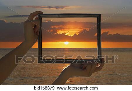 Stock Photo - Caucasian woman holding frame over dramatic sunset sky and ocean. Fotosearch
