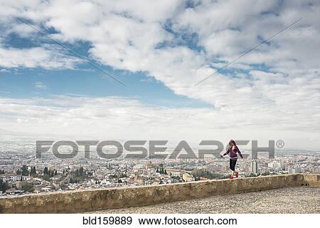 Stock Photo - Caucasian woman on wall overlooking scenic view of cityscape, Granada, Spain. Fotosearch