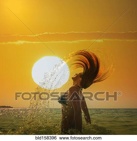 Stock Photograph - Caucasian woman tossing hair in ocean under sunset sky. Fotosearch