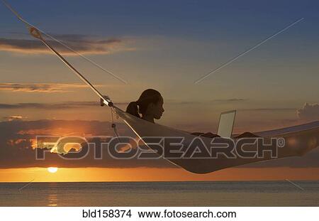 Picture - Caucasian woman using laptop in hammock near ocean at sunset. Fotosearch