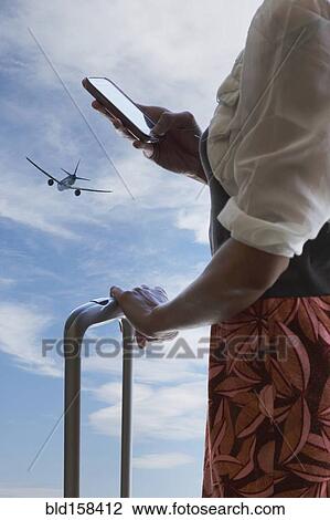 Stock Image - Chinese traveler using cell phone under airplane in blue sky. Fotosearch