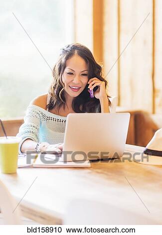 Stock Image - Chinese woman on cell phone using laptop in cafe. Fotosearch