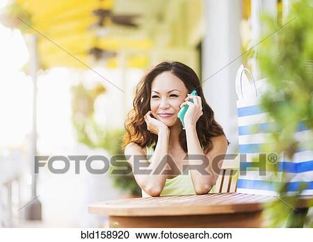 Stock Image - Chinese woman talking on cell phone at sidewalk cafe. Fotosearch